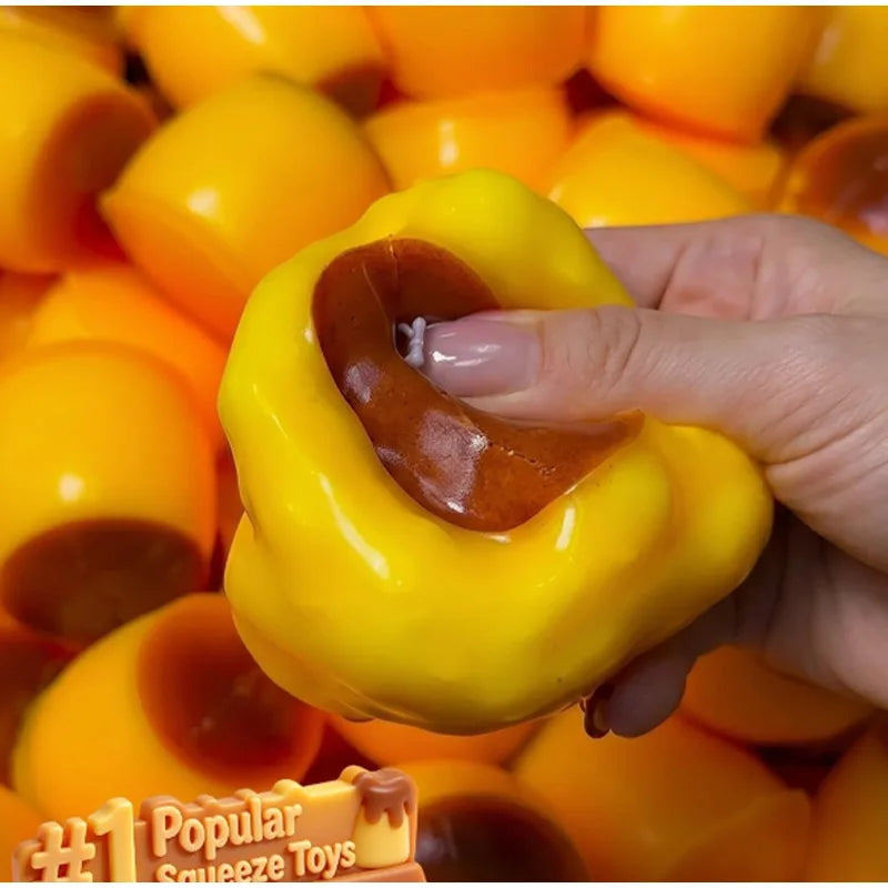 A hand holds a bright yellow caramel pudding squishy stress relief ball surrounded by other similar toys.