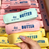 A hand holds a pink squeeze toy butter block next to a stack of similar butter blocks.