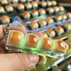 A hand holds a clear LED mechanical keycap fidget keychain with glowing green lights, positioned above a tray of similar item