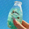 A hand holds a clear, light green bottle filled with liquid and a small, squishy toy inside.