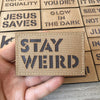 Une main tient un écusson tactique marron avec l'inscription STAY WEIRD, parmi d'autres écussons.