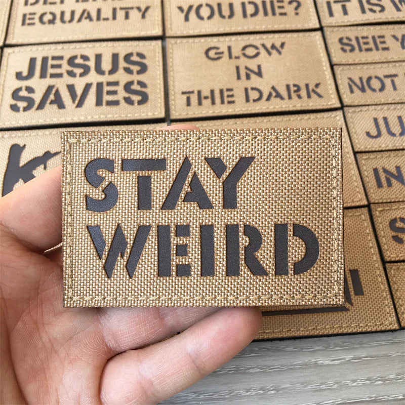 Une main tient un écusson tactique marron avec l'inscription STAY WEIRD, parmi d'autres écussons.