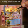 A hand is applying detail to a pink and blue wooden miniature candy shop diorama kit.