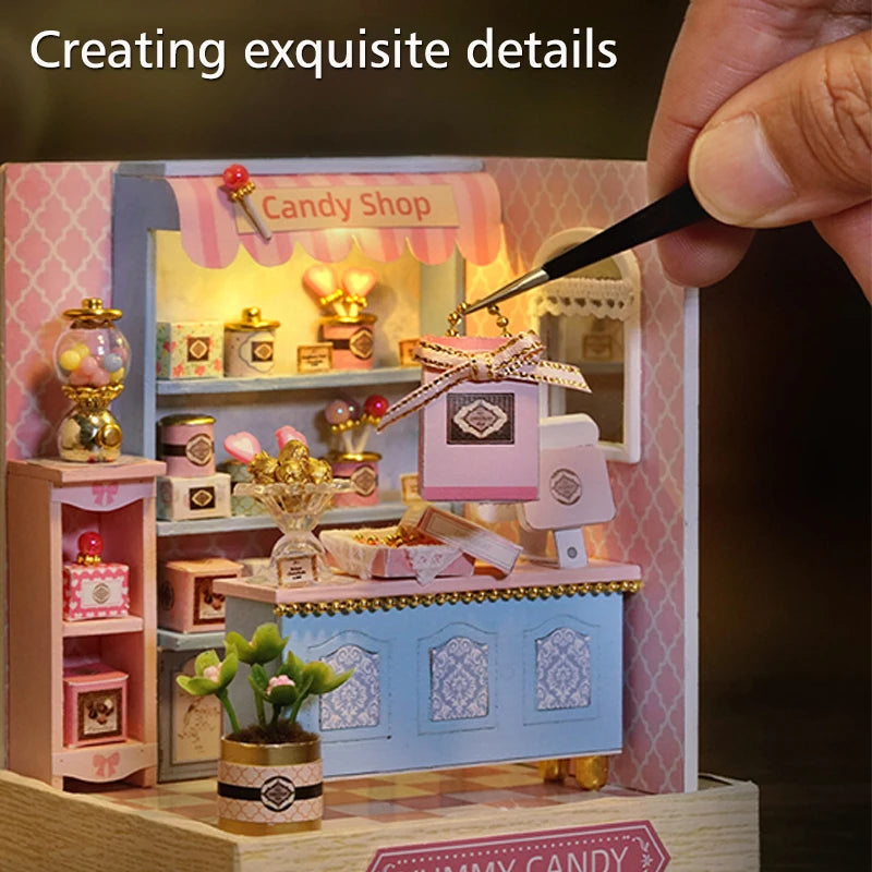 A hand is applying detail to a pink and blue wooden miniature candy shop diorama kit.