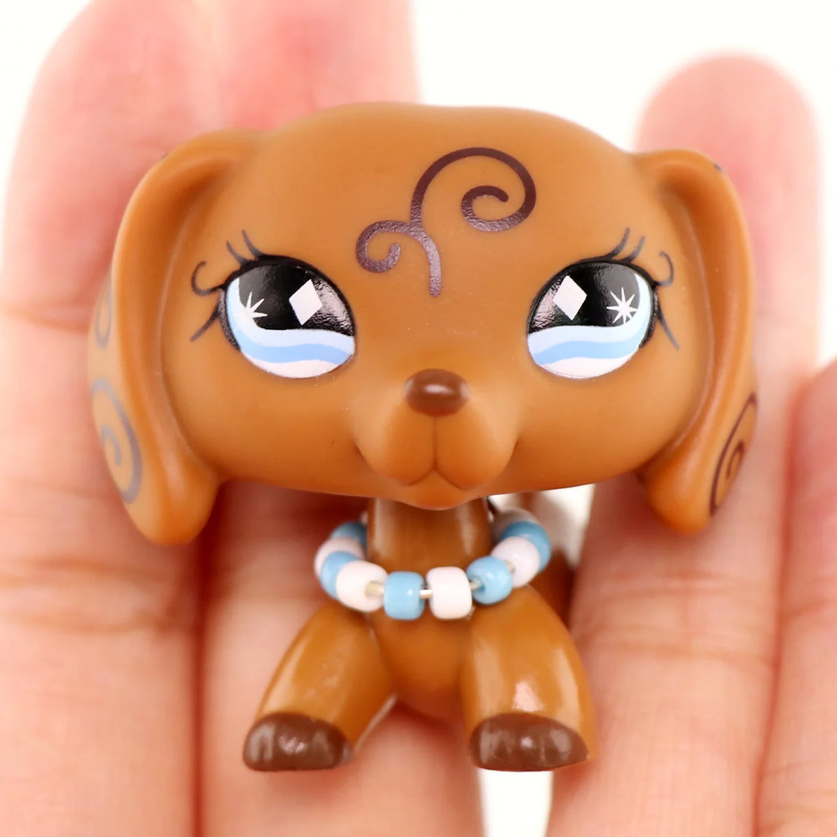 A small, tan-colored miniature dog figure with large eyes and a blue beaded collar is held in a person's hand.