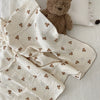 A white muslin swaddle blanket with a floral print is draped next to a brown teddy bear on a white surface.