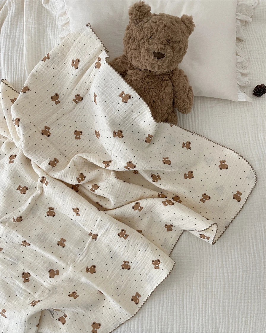 A white muslin swaddle blanket with a floral print is draped next to a brown teddy bear on a white surface.