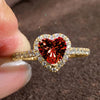 A close-up of a gold heart-shaped ring featuring a large red gemstone surrounded by pave diamonds.