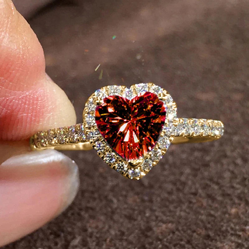 A close-up of a gold heart-shaped ring featuring a large red gemstone surrounded by pave diamonds.