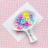 A white spoon holds colorful pastel wavy acrylic beads with floral edges on a pink checkered background.