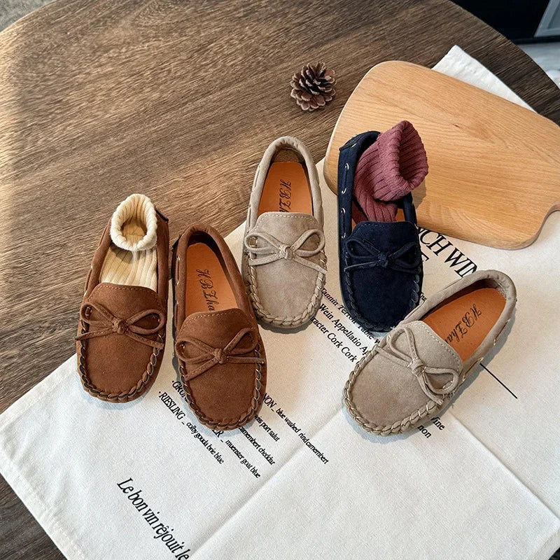 A pair of girls' brown moccasins with bow details and suede accents resting on a white cloth.