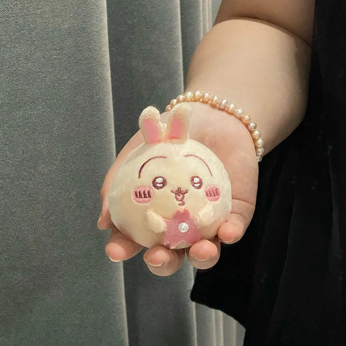 A small, round, light pink plush charm shaped like a rabbit with pink accents is held in a hand wearing a pearl bracelet.