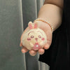A small, round, light pink plush charm shaped like a rabbit with pink accents is held in a hand wearing a pearl bracelet.