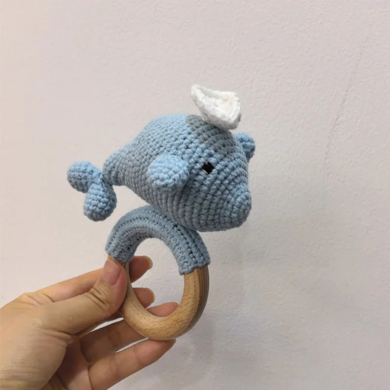 A handmade blue otter rattle and teether ring made of wood is held in a hand against a white background.
