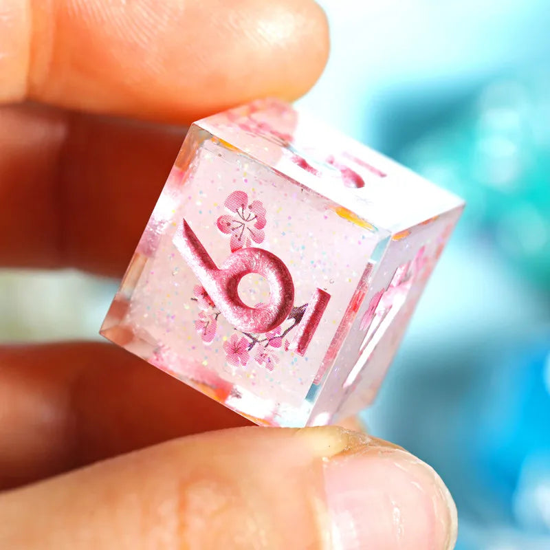 A hand holds a pink resin polyhedral die featuring a floral design and glitter.
