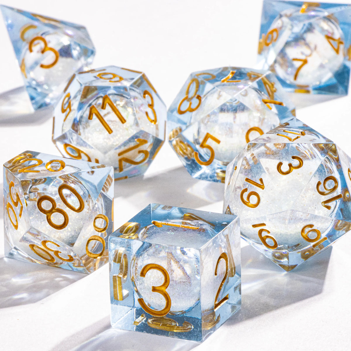 A collection of clear blue liquid core polyhedral dice with gold galaxy finish scattered on a white surface.
