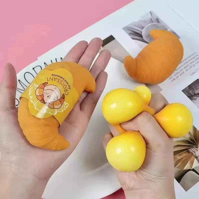 A hand holds an orange squishy hamburger stress ball and a yellow squishy ball against a light background.
