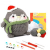 A gray crocheted penguin toy with a green scarf and a DIY kit box containing yarn and tools.