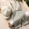 A set of a light green knit romper and matching newsboy cap laid on a beige woven background.