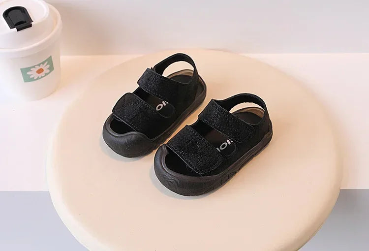 A pair of black toddler soft-soled sandals for boys and girls resting on a light beige surface.