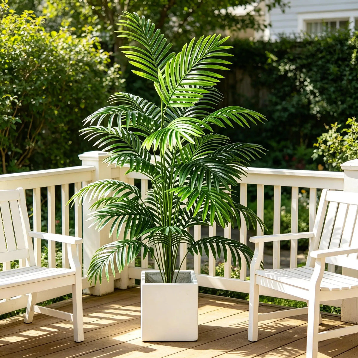 Artificial areca palm plant in a white decorative pot sitting on a wooden porch with white chairs.