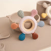 A pink silicone teether shaped like a rudder with a wooden ring and various colored silicone protrusions.