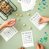 Hands are preparing a bingo game with paper sheets and bingo balls on a green surface.