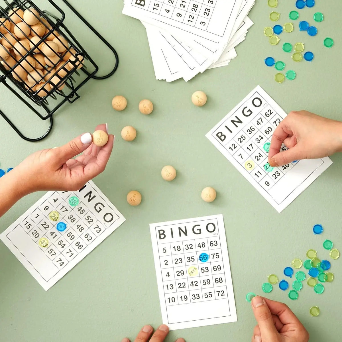 Hands are preparing a bingo game with paper sheets and bingo balls on a green surface.