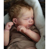 A close-up photo of a newborn reborn baby doll with rooted hair sleeping, wrapped in a soft fabric.
