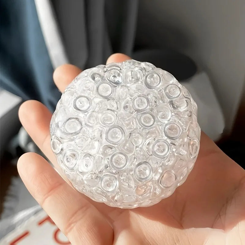 A hand holds a clear, spherical fidget ball made of soft, squeezable bubbles for stress relief.