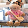 A soft plush cow doll with white and black markings, wearing a pink sweater and carrying a tan bag.