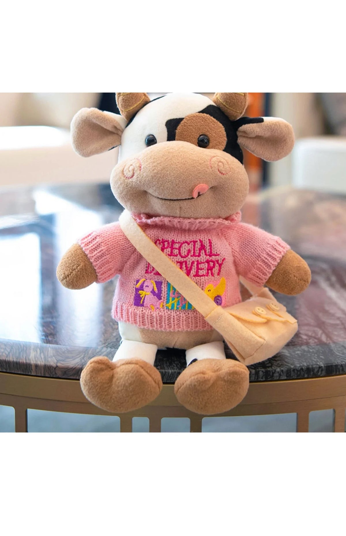 A soft plush cow doll with white and black markings, wearing a pink sweater and carrying a tan bag.