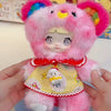 A pink fluffy collectible doll with large eyes and a yellow garment is held in a person's hands.