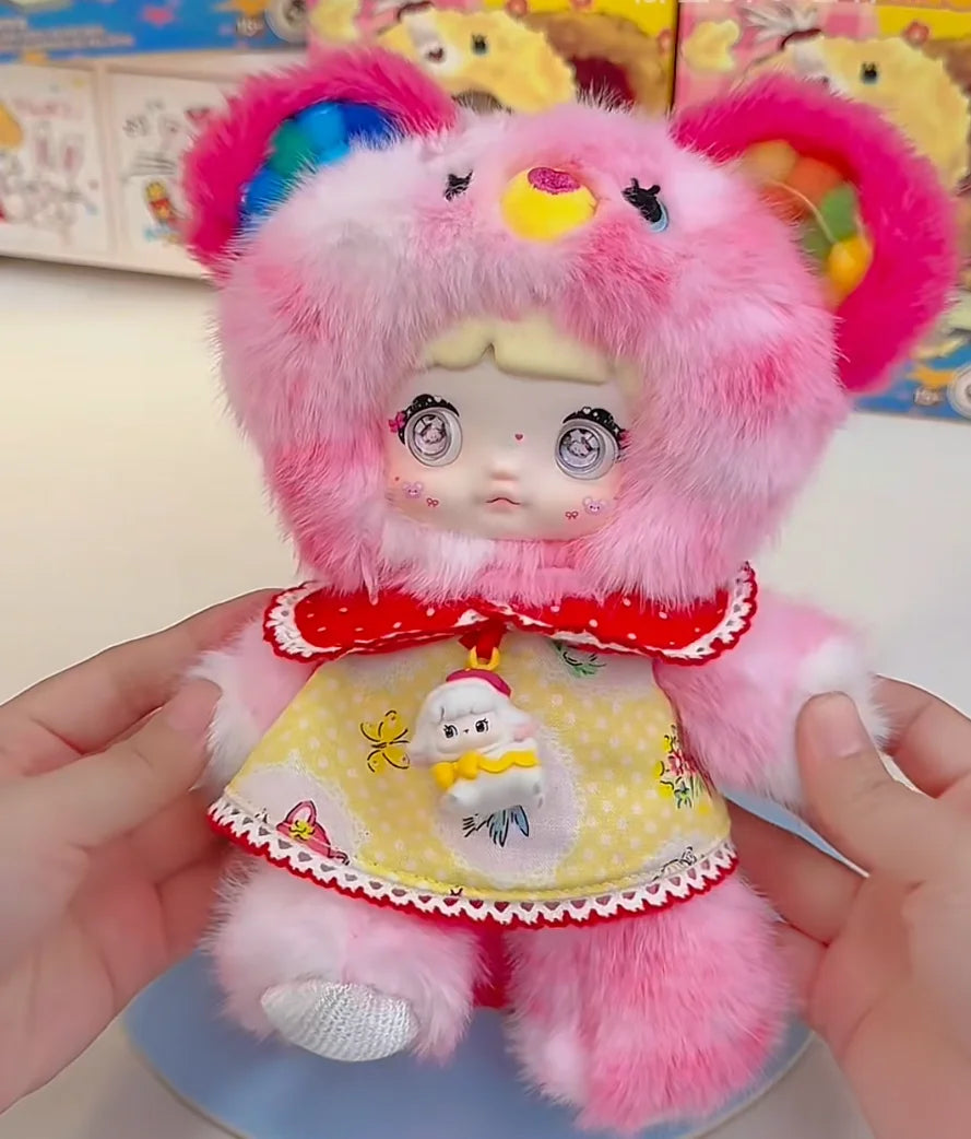 A pink fluffy collectible doll with large eyes and a yellow garment is held in a person's hands.