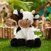 A white and black cow plush toy with a brown nose lying on green grass surrounded by wood logs.