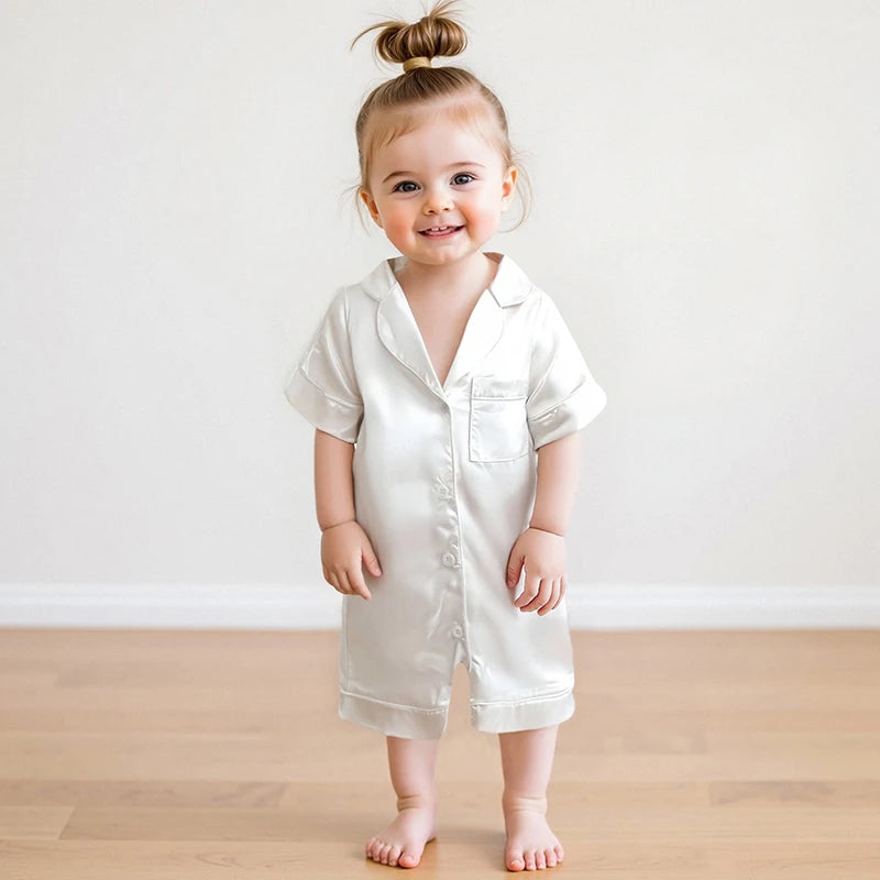 A toddler girl wearing a light satin button-up romper sleepwear standing against a white background.