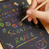 A hand holds a pencil drawing colorful scratch art on a black notebook page.