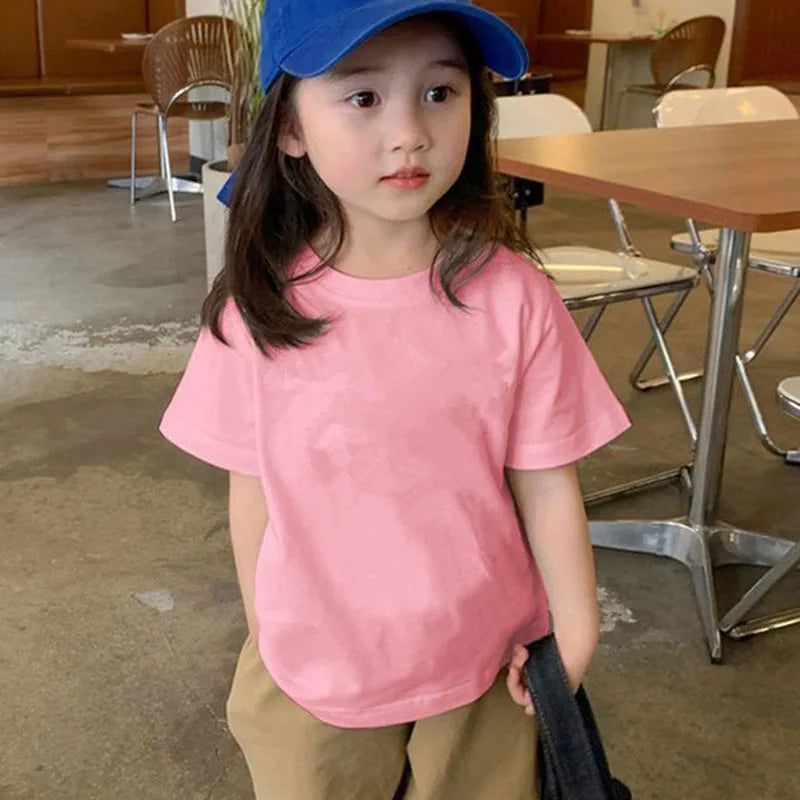 A young girl wearing a pink classic cotton crew neck t-shirt and a blue baseball cap standing indoors.
