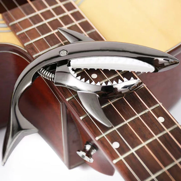 A close-up of a metal shark guitar capo clamped onto the fretboard of an acoustic or electric guitar.