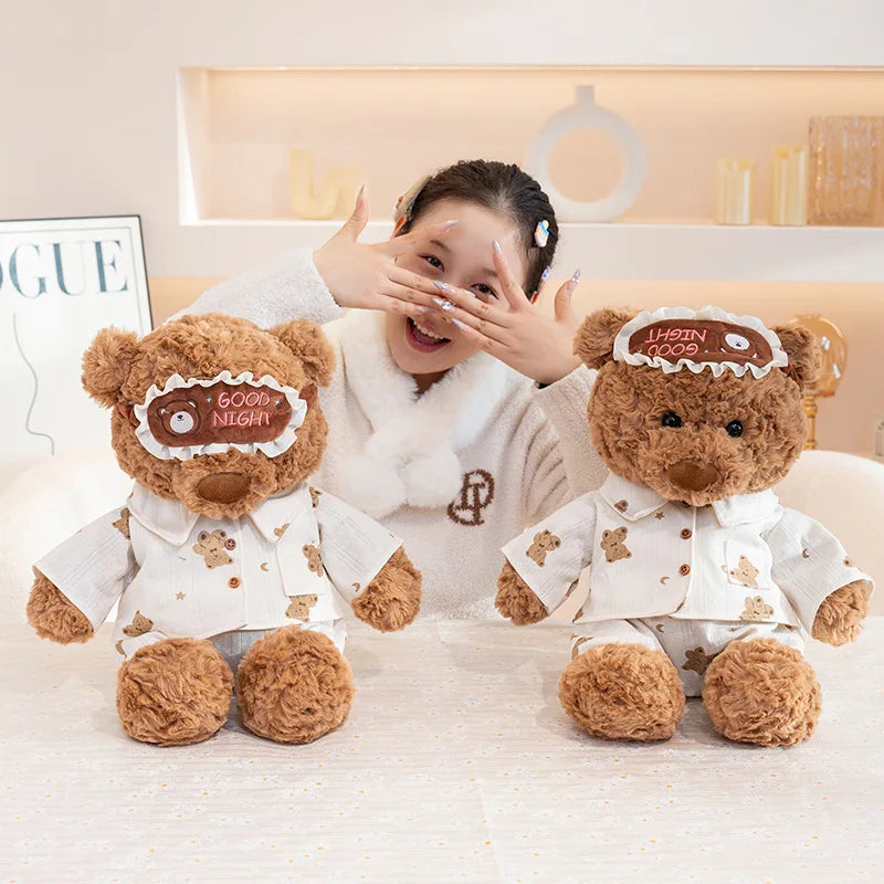 A smiling girl is playfully covering her eyes with her hand while sitting next to a brown teddy bear plush toy.