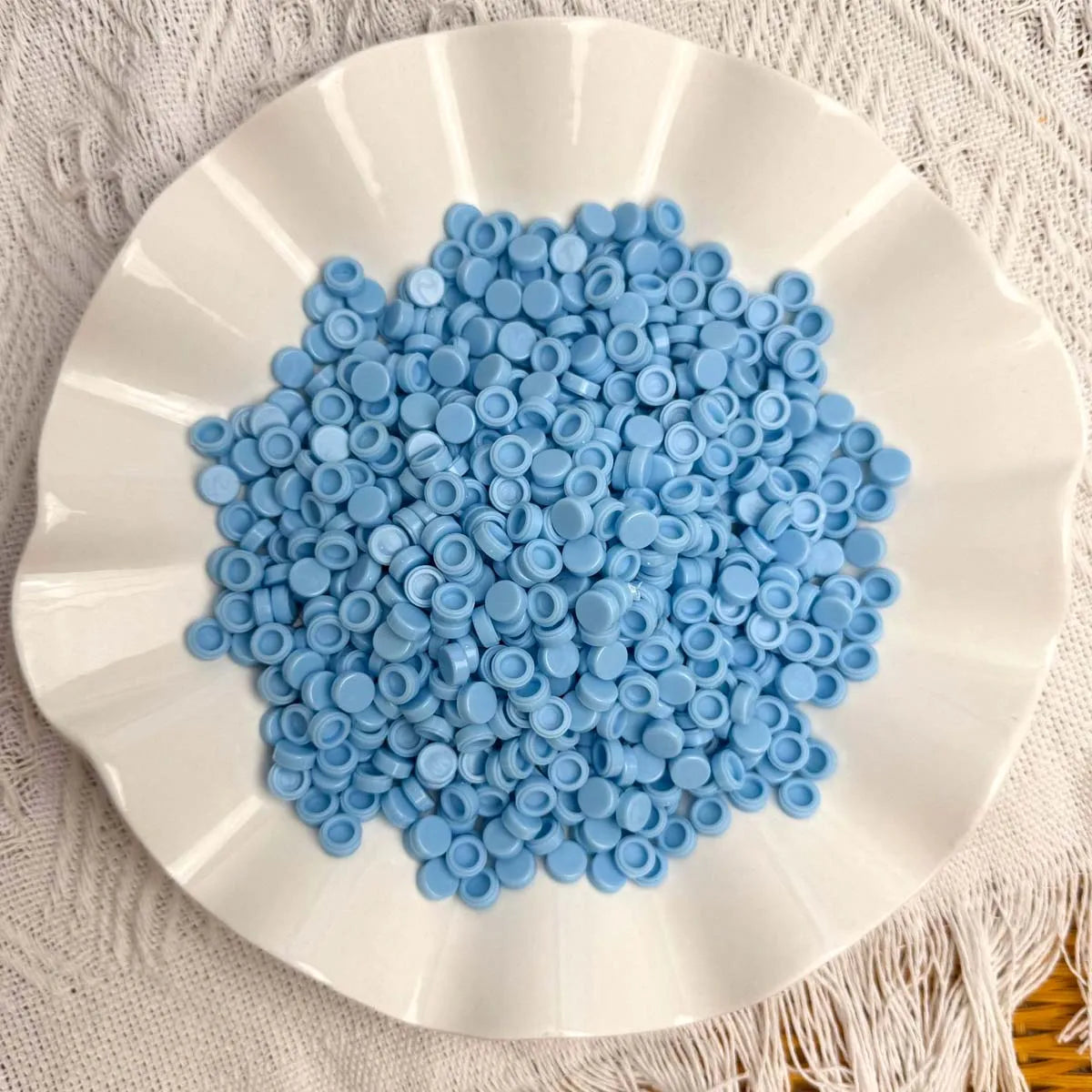 A white plate filled with numerous small, light blue plastic beads for crafting.