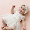 A baby girl wearing a white lace tulle dress and matching hat lying on a pink surface.
