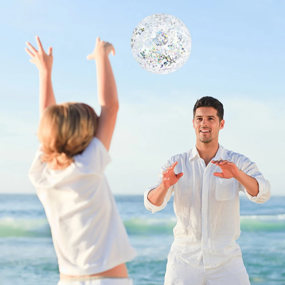 A man and a woman are playing with a colorful sequined inflatable beach ball near the ocean.