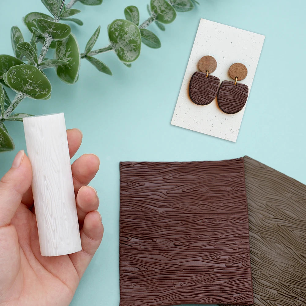 A hand holds a white floral texture roller next to a piece of dark brown wood and a small gold-colored jewelry display.