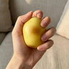A hand is holding a yellow fluid potato squishy toy against a soft, light-colored background.