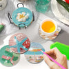 A hand holds a pink brush over several finished diamond painting coasters and a green tool on a white surface.