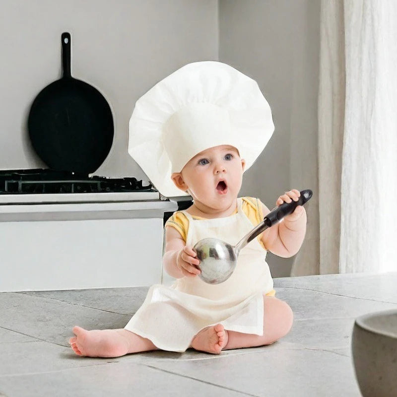 A baby wearing a white chef hat and apron sits on the floor in a kitchen setting with a black pan visible.