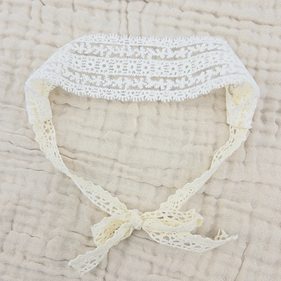 White floral lace baby headband with a gathered bow on a beige textured fabric.