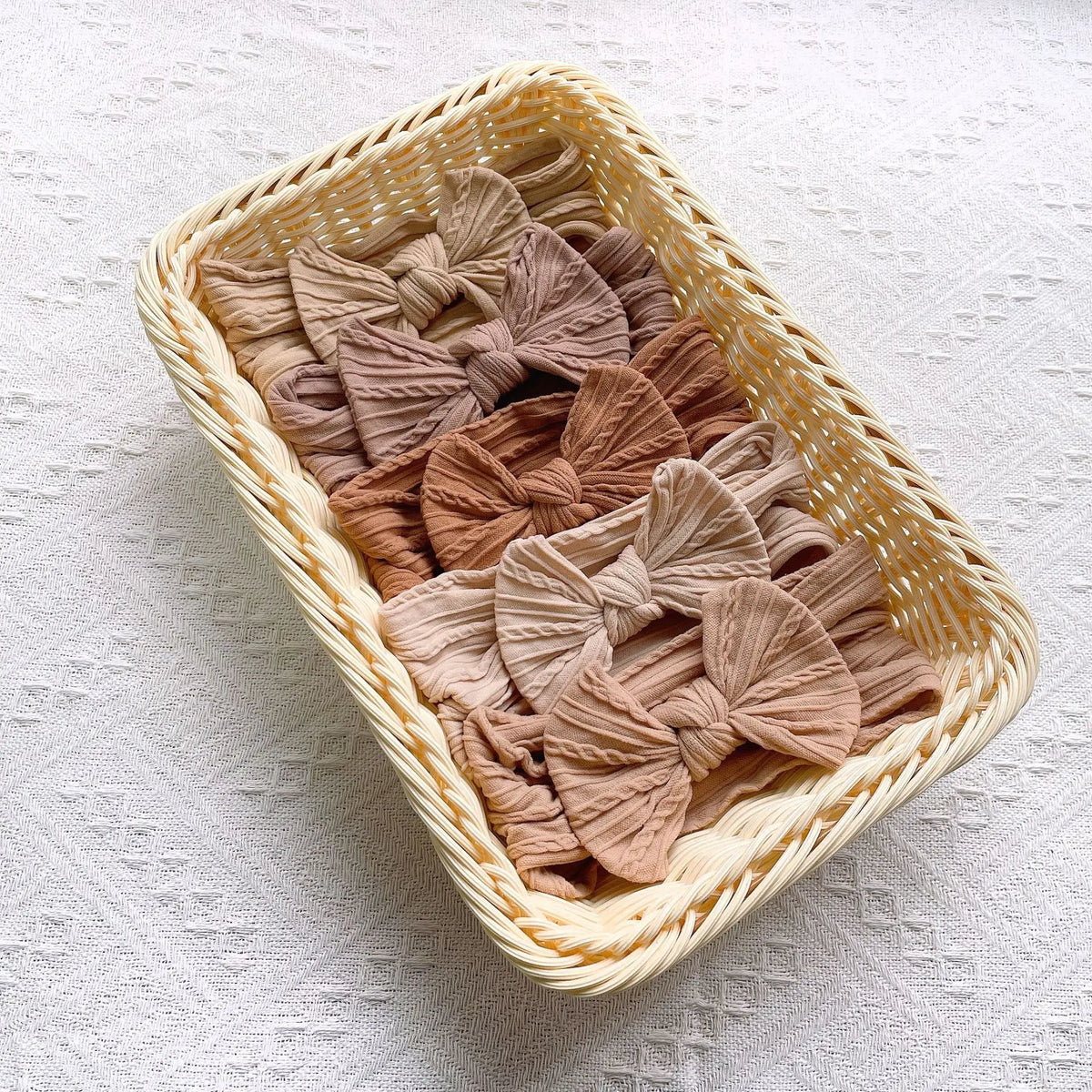 A woven basket filled with five light brown fabric bow baby headbands on a white textured surface.