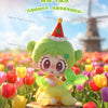 A cute green figurine of a puppy with a red party hat stands in a field of colorful flowers.